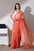 A woman models a peach-orange and red tie-dye georgette saree with a white sleeveless blouse, standing against a textured beige wall.