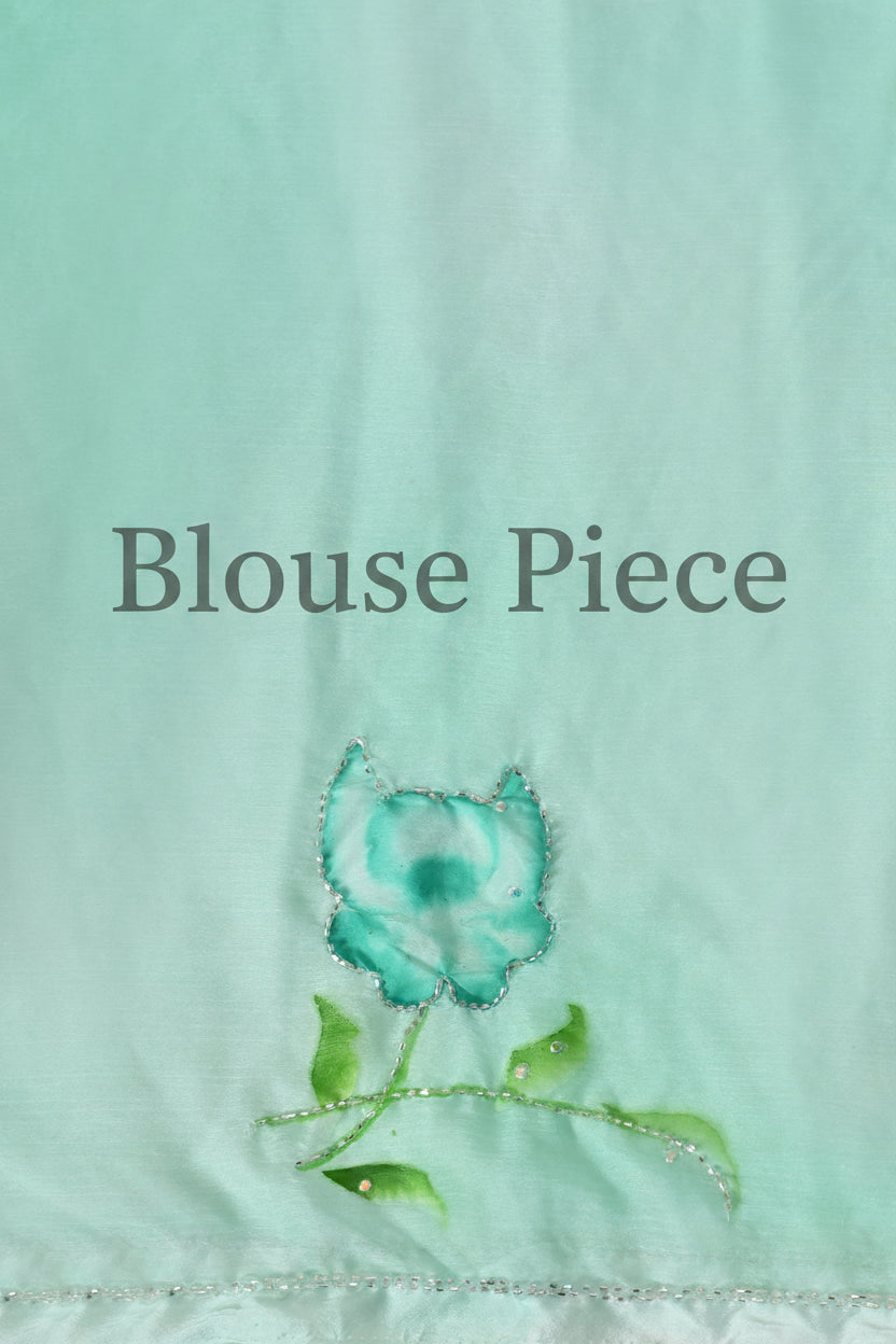 A close-up of a mint green fabric surface featuring a small embroidered flower motif in turquoise and green with subtle shimmering details.